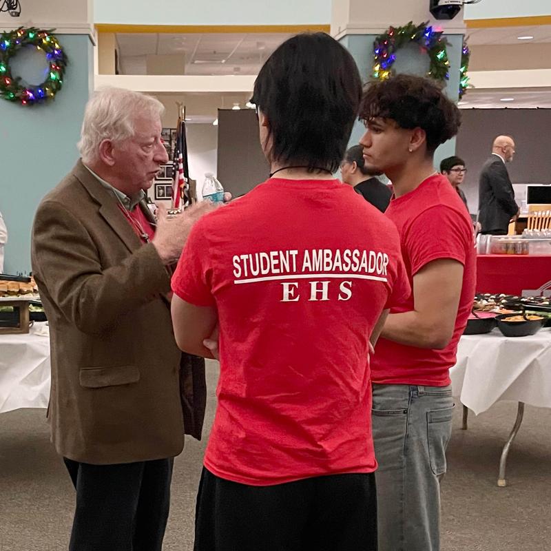 A group of people, including two students in red shirts, engage in conversation at an event.