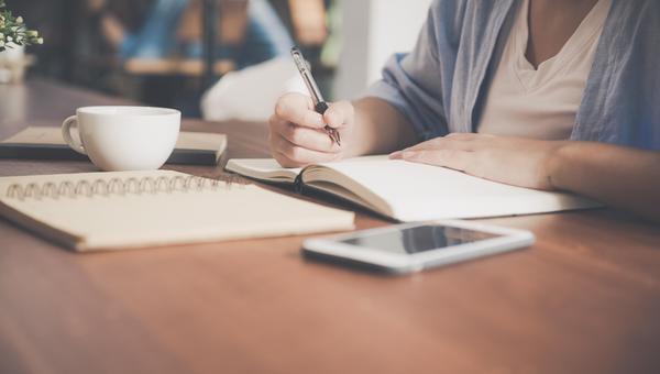 Person writing in a notebook with a cup on the table.
