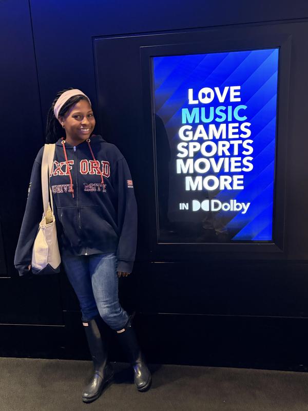 Girl in a hoodie poses beside a vibrant Dolby advertisement.
