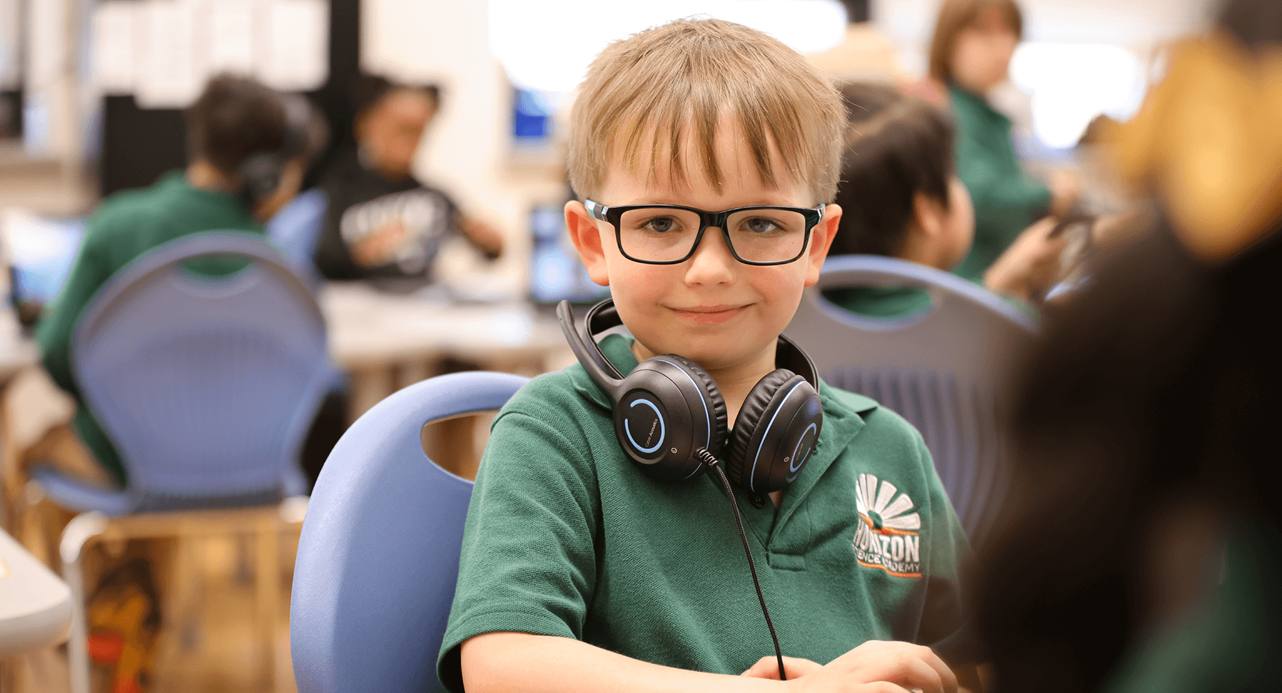 Boy with glasses and headphones smiling in a classroom setting.