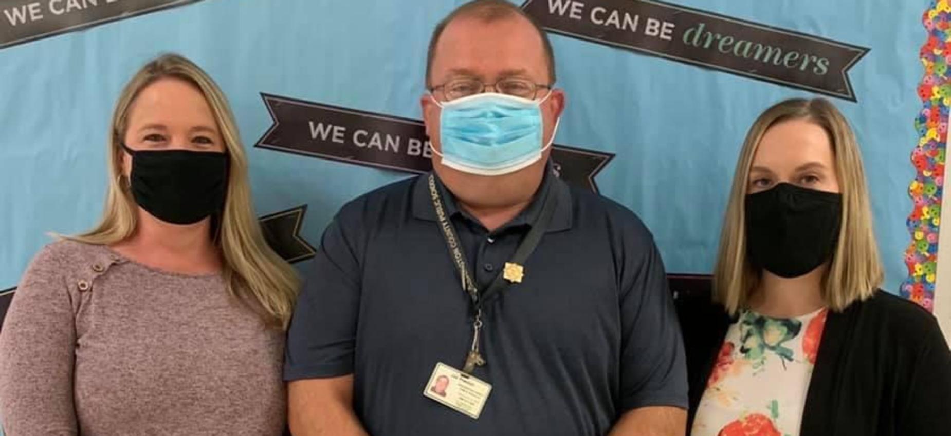 Three teachers wear masks in front of a bulletin board.