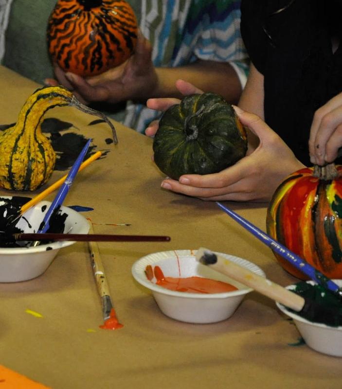 Hands holding decorated pumpkins and gourds with paint and brushes on a table.