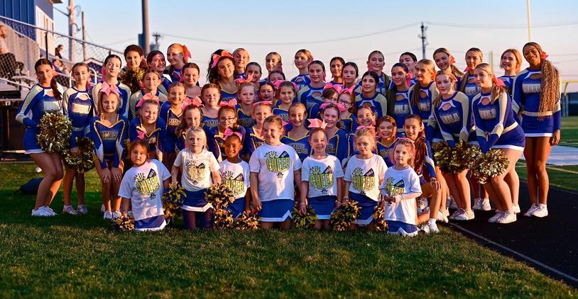 A team of cheerleaders, both young and older, pose together in their uniforms on a field.