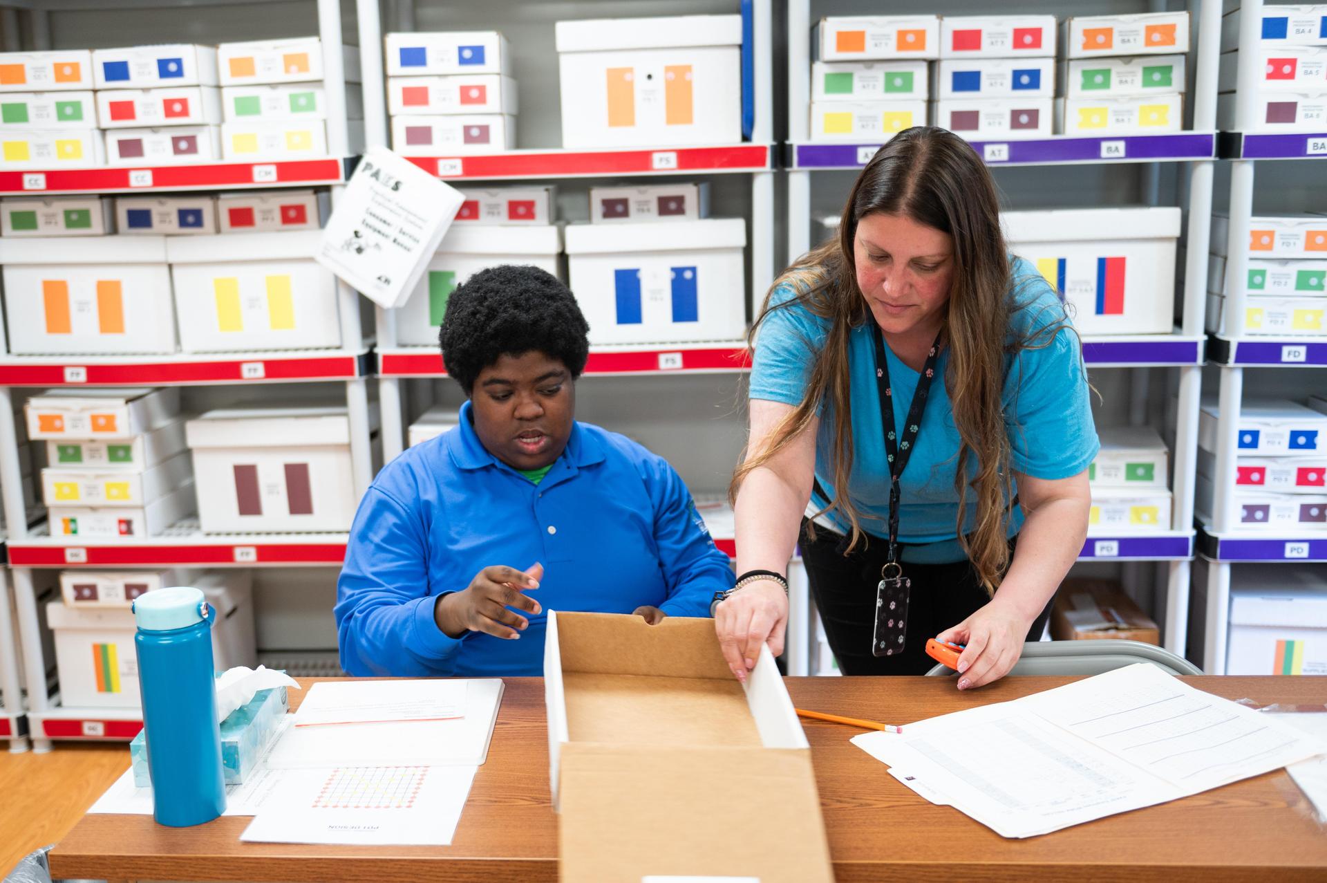 Student sitting at table on left with teacher on right giving instructions.