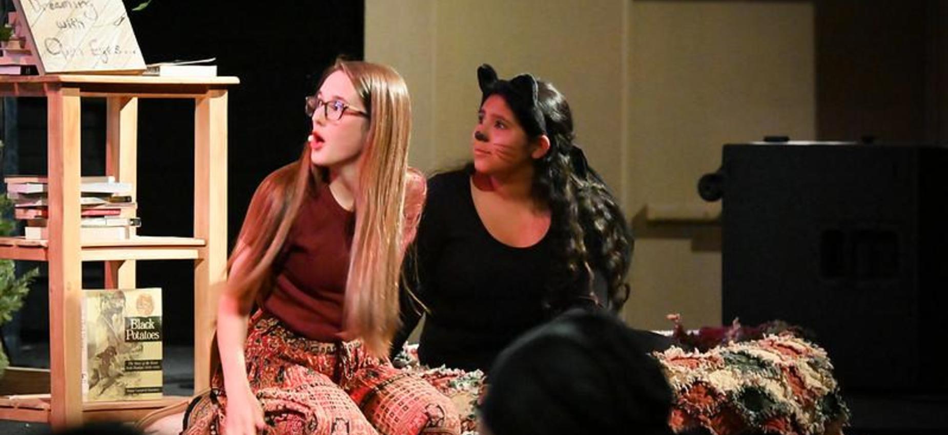 Two girls sitting on a stage, one wearing cat ears, engaged in a performance.