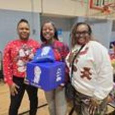 Three women in festive sweaters smiling while holding a blue box.