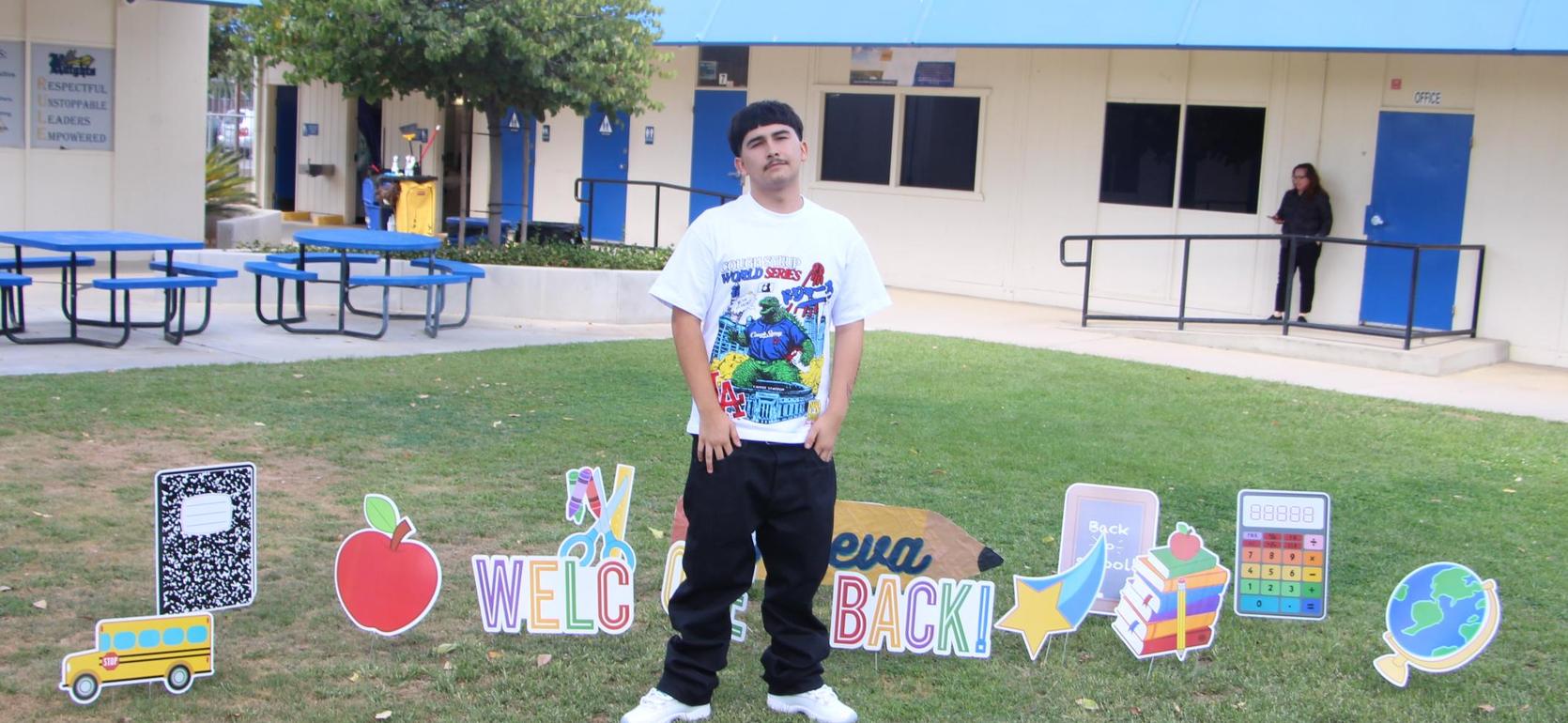 A person in a white t-shirt stands in front of colorful welcome signs on grass.