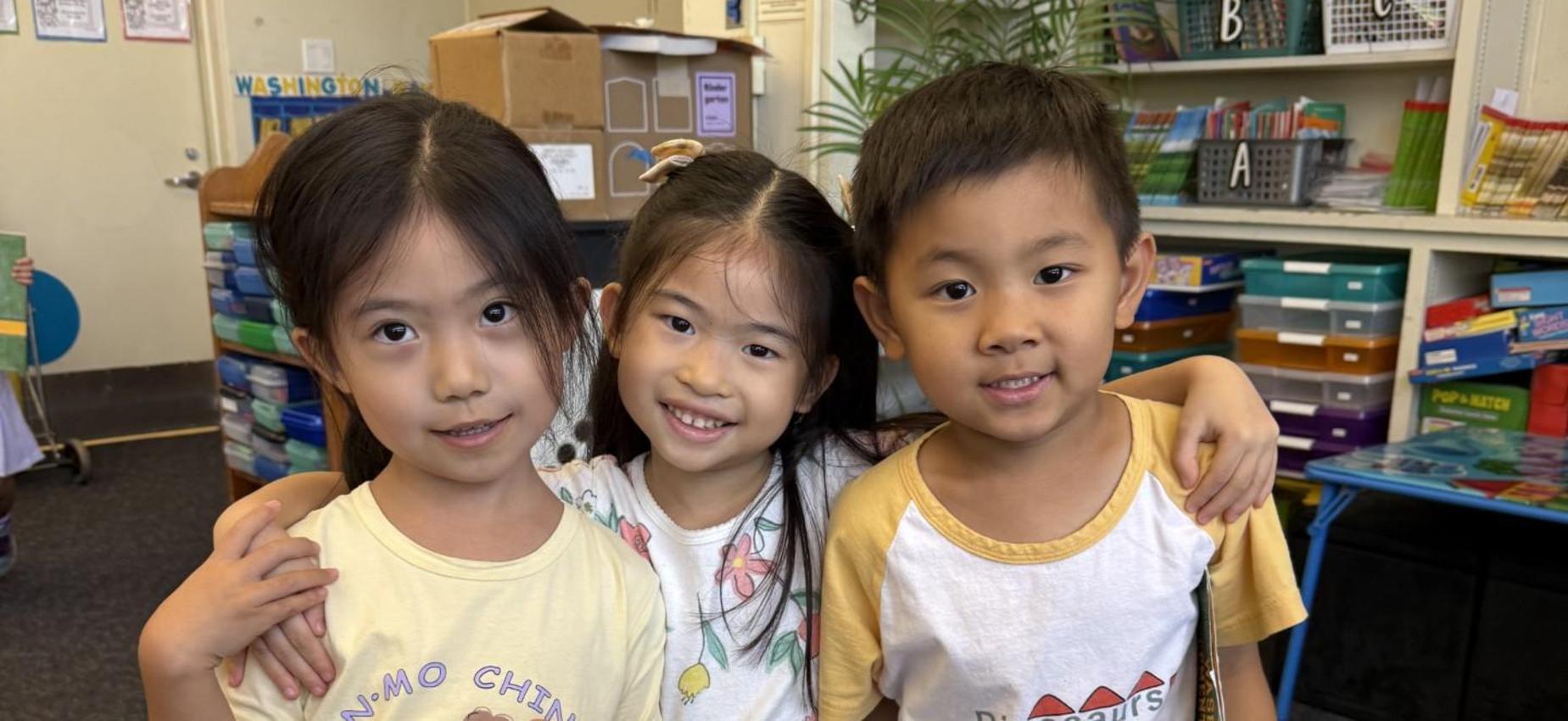 Three children posing together, smiling in a classroom setting.