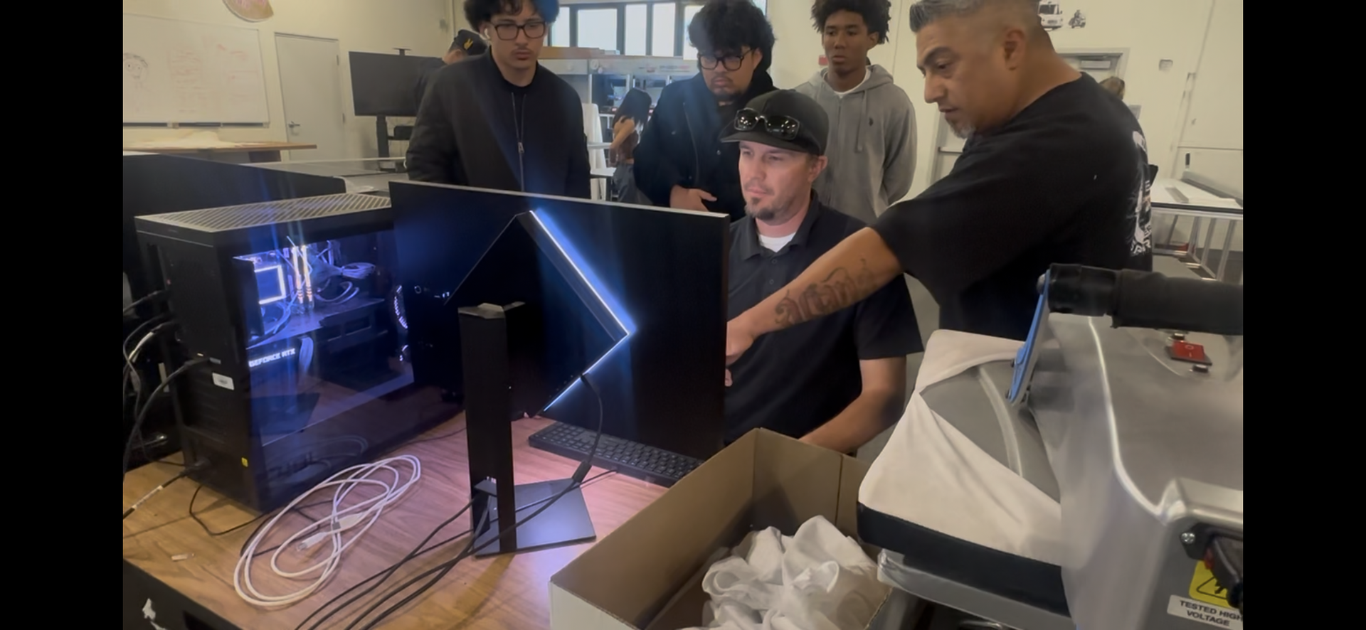 person sitting at a computer with another person pointing at the screen and 3 other people standing behind the person sitting at the computer