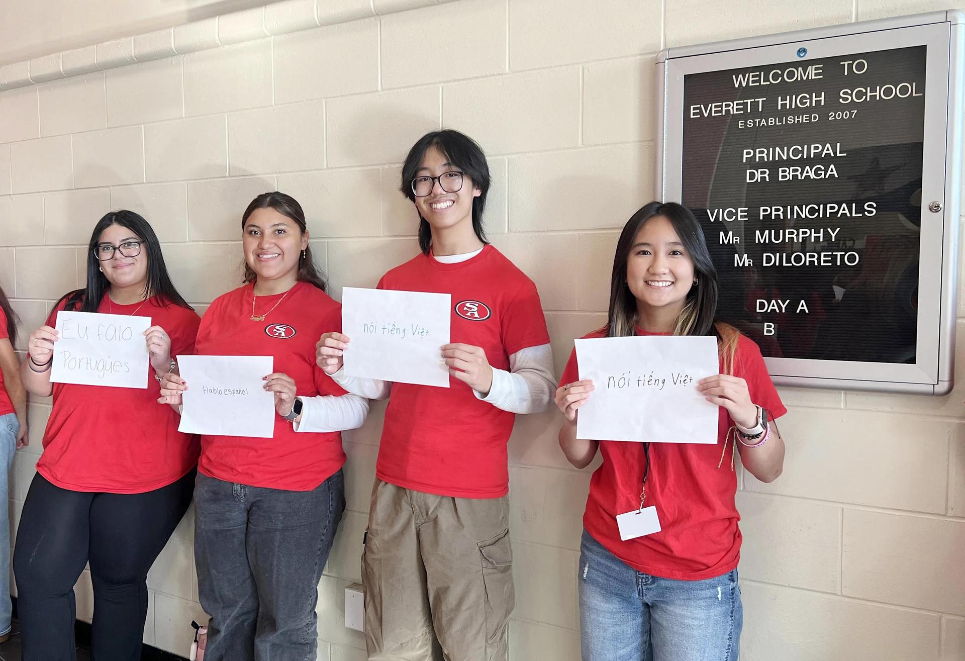 Students in red shirts holding signs in front of a school welcome board.