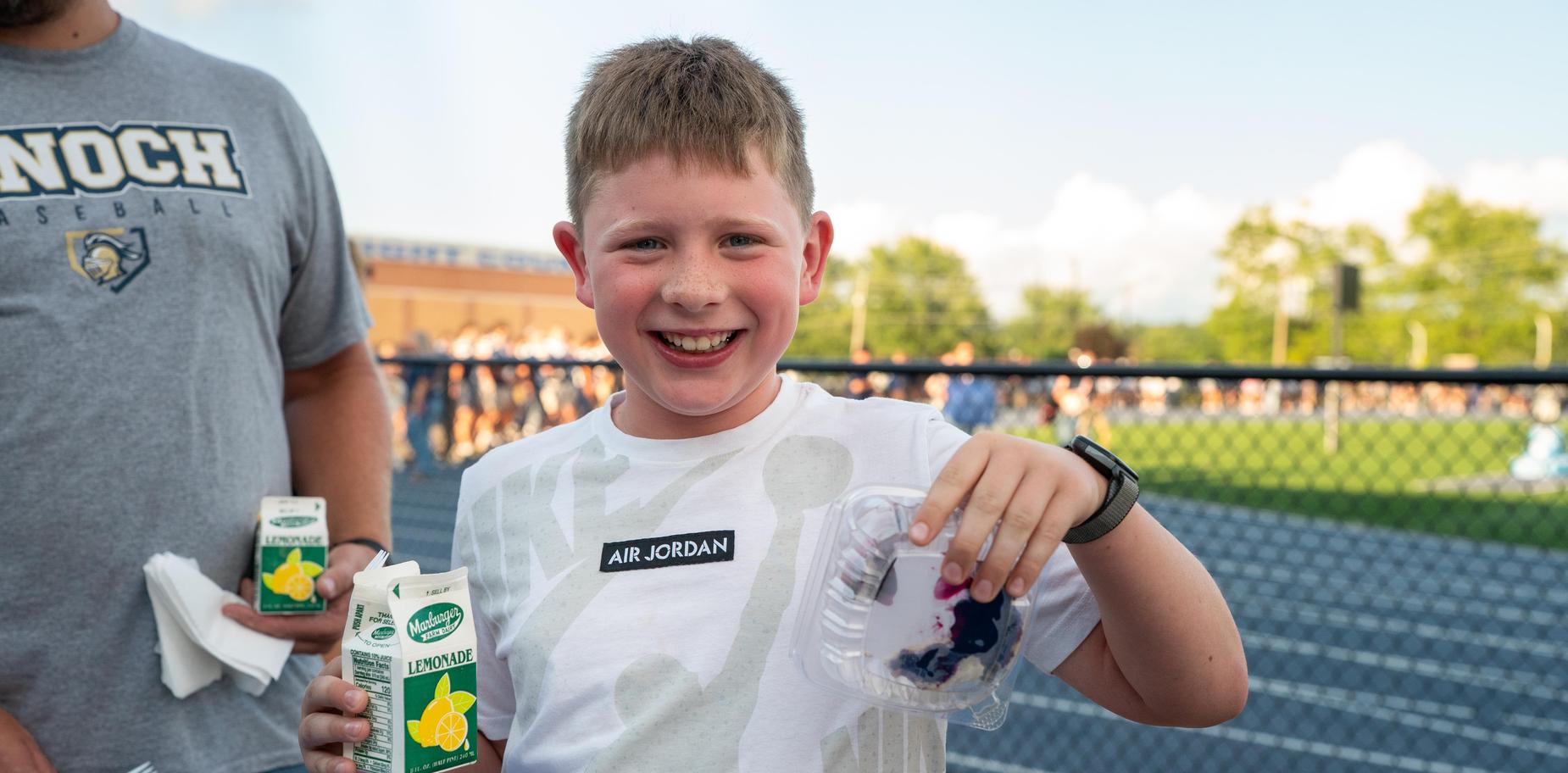 Smiling boy holding a snack and drink at a sports event with a cheering crowd.