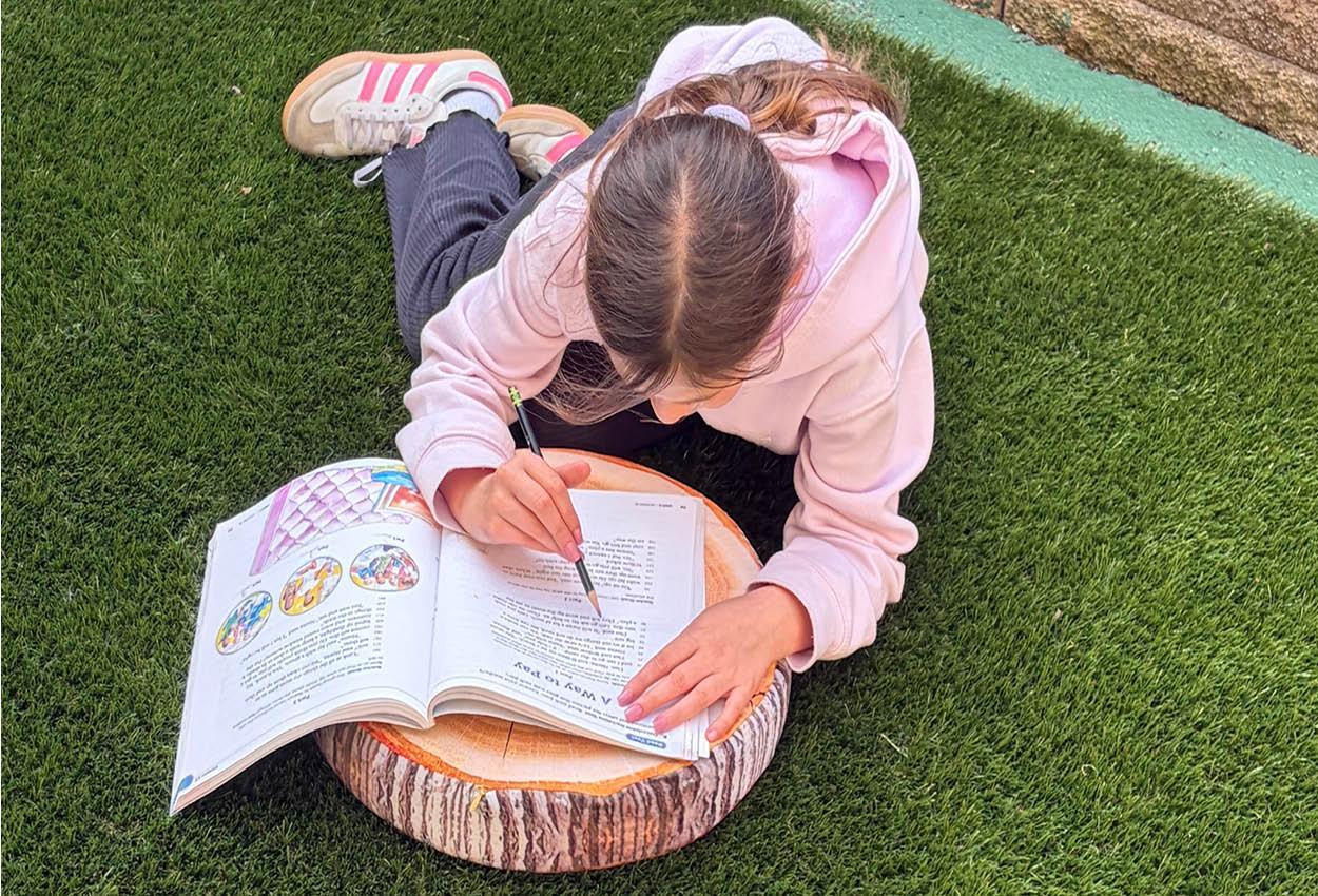 Child writing in a book on a wooden log seat, surrounded by green grass.