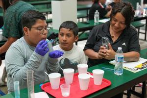 a boy blows bubbles into his had while another boy watches and a lady takes a photo