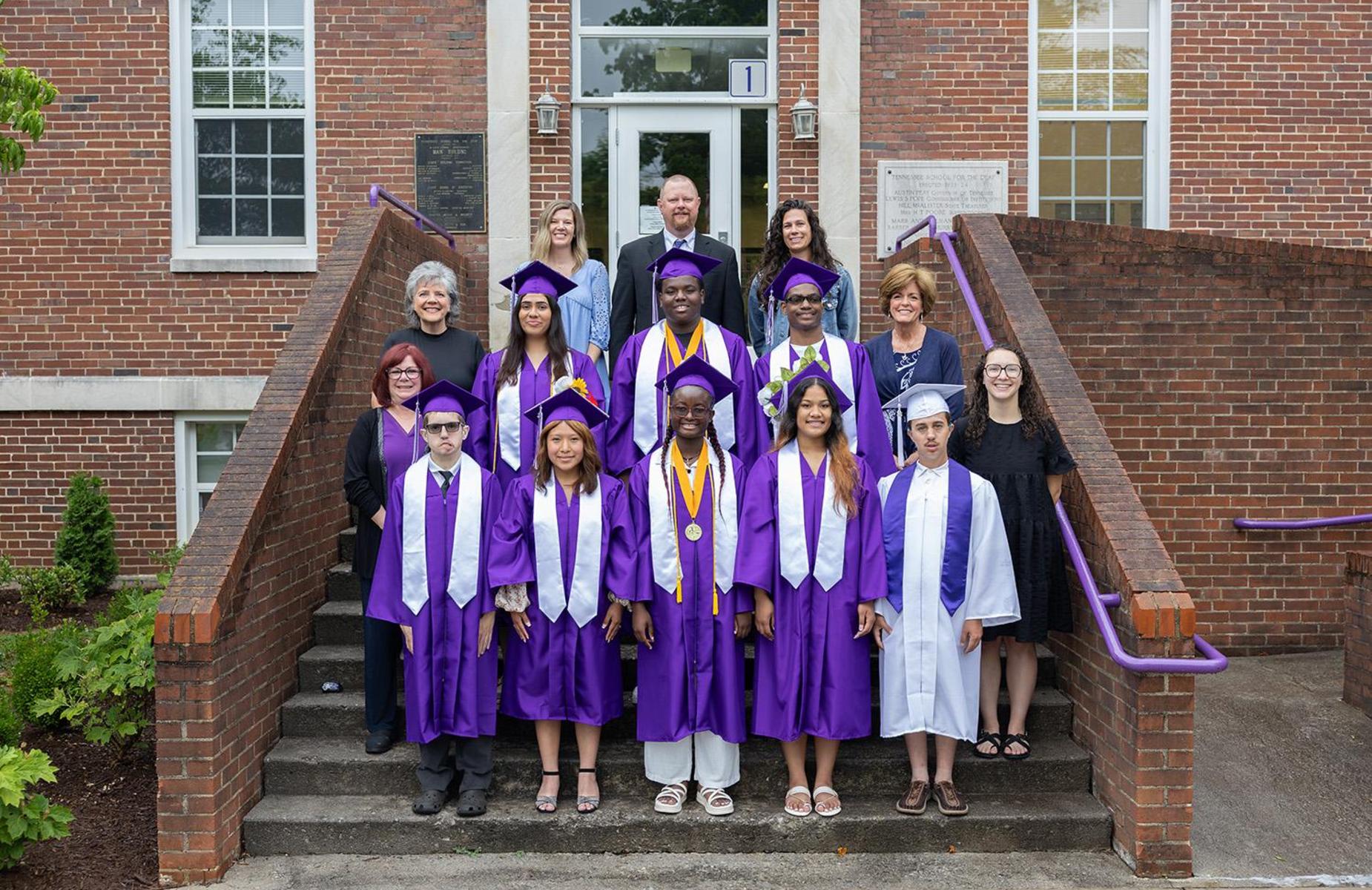 Group of graduates in purple gowns posing on stairs outside a school building.