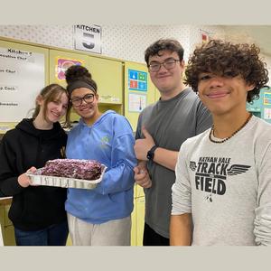 Students holding a chocolate dessert in a kitchen environment.