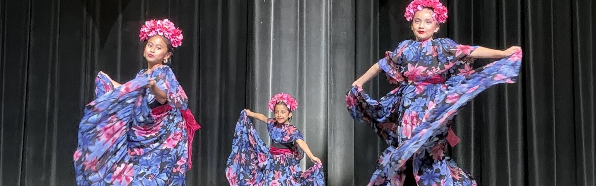 Young dancers in floral dresses perform on stage, showcasing traditional movements.