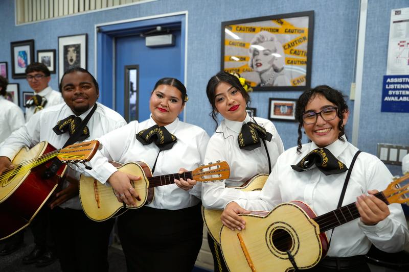 Four students from Santa Fe High School stand together, dressed in traditional mariachi outfits, holding up their guitars and smiling at the camera inside a district board room after a performance.