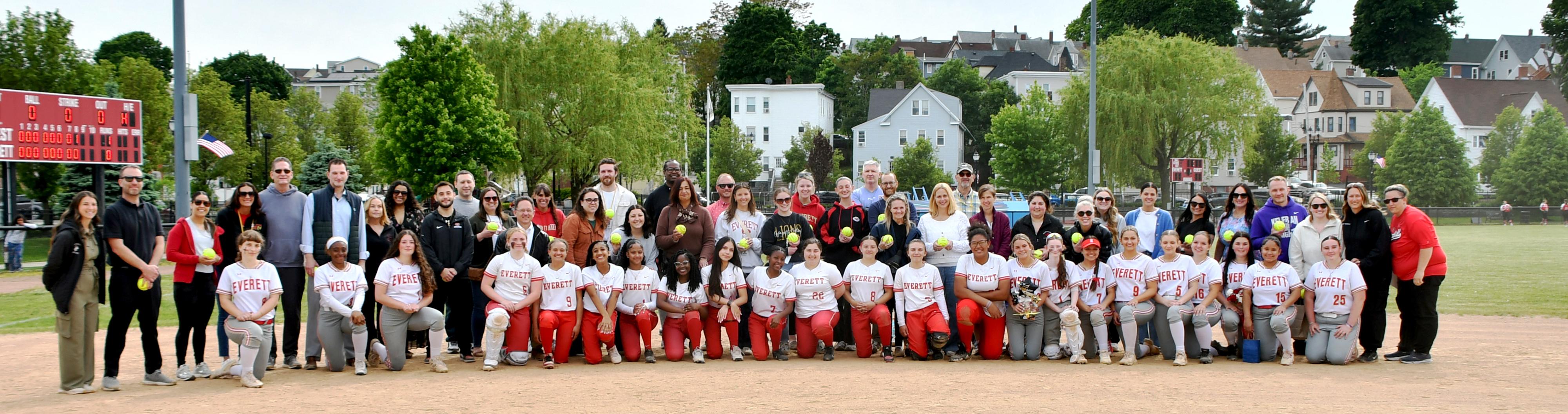 A large group of students, families, and staff pose on a softball field.