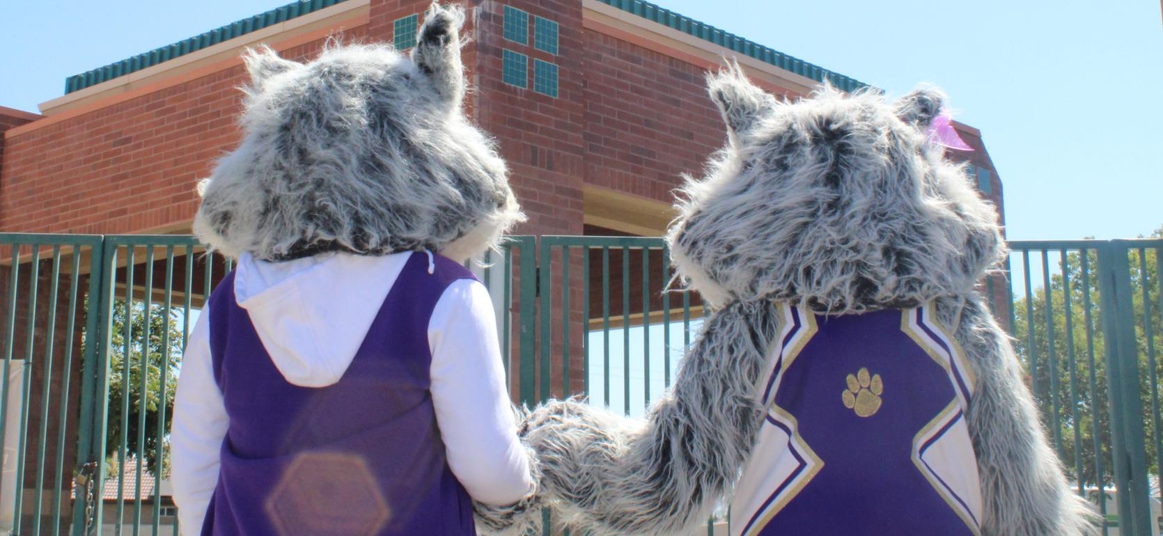 Two costumed mascots holding hands, facing away from the camera outside a building.