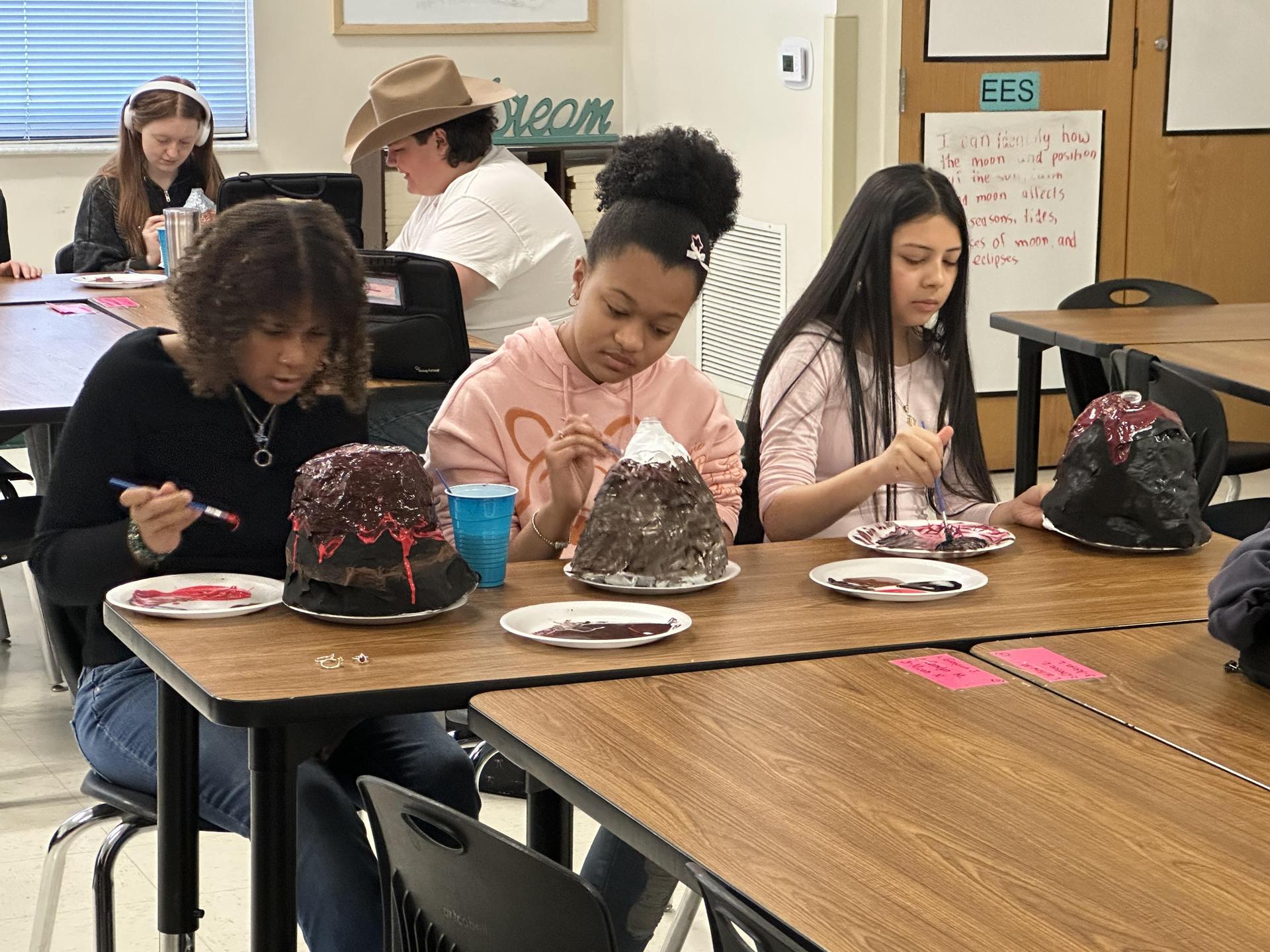 Three students paint different volcano models at a table.