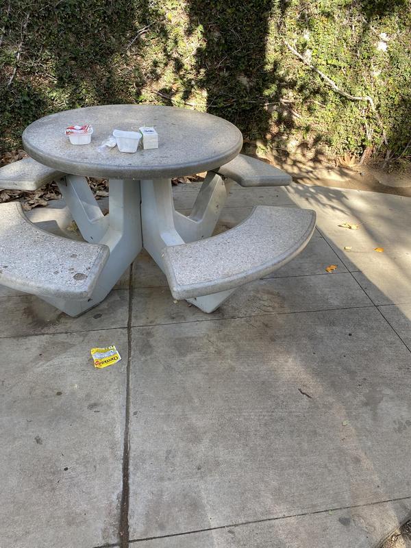 A concrete picnic table with a round top and four attached curved bench seats on a patio. Trash including napkins and containers are placed on the table surface, with a yellow cereal on the ground in front.