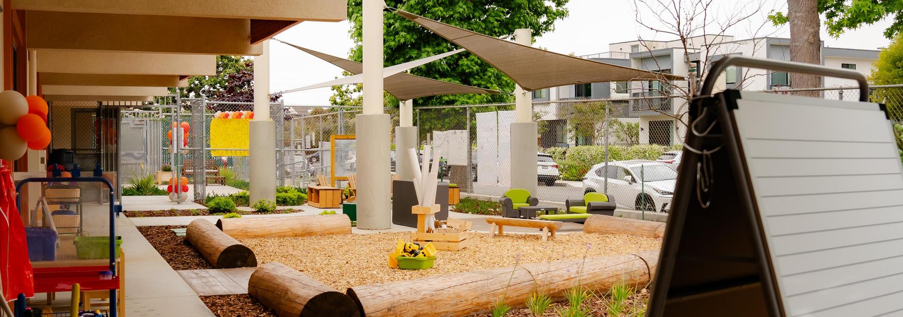 Preschool outdoor area with logs, woodchips, shade sails with trees in the background