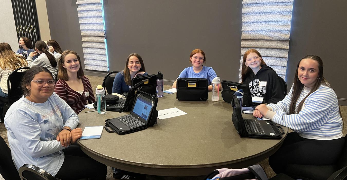 Six individuals sitting at a round table, engaged in a group discussion with laptops.