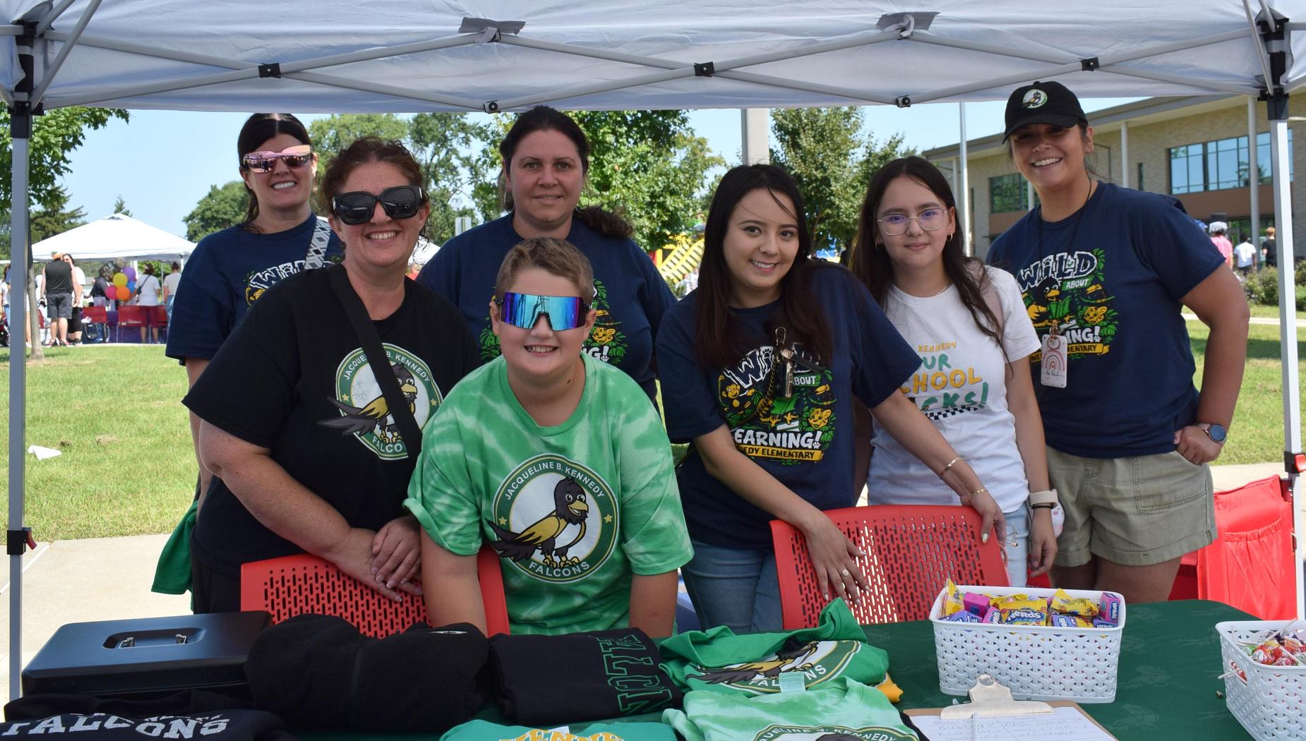 Seven women and one boy at a booth with merchandise, smiling and engaged with attendees.