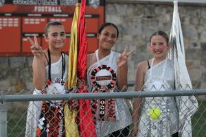 Three girls in cheerleading outfits holding flags, smiling and making peace signs.