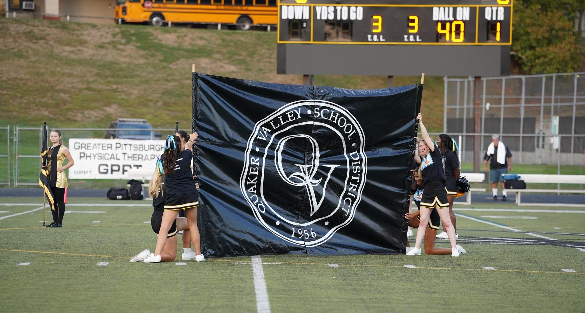 Cheerleaders holding a large school banner on a football field with a scoreboard.