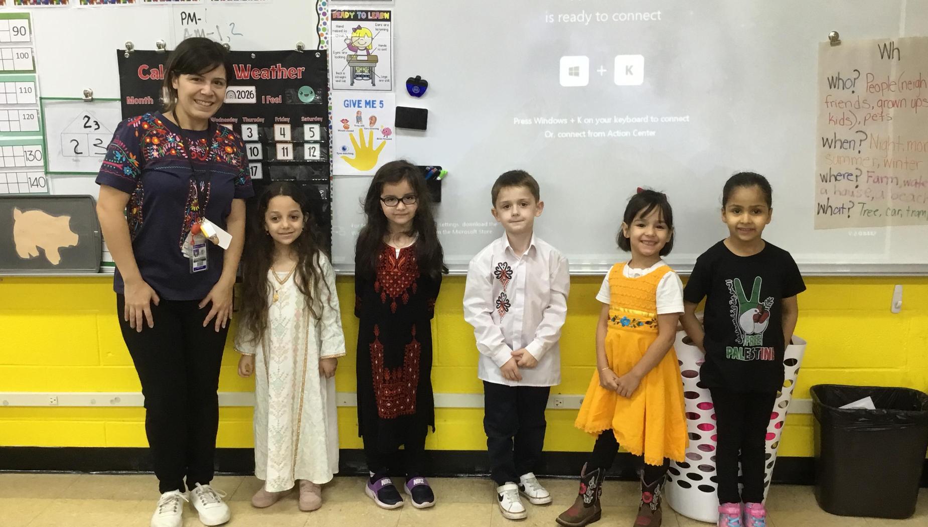 Six children in the classroom with a teacher, smiling together in front of a display screen.