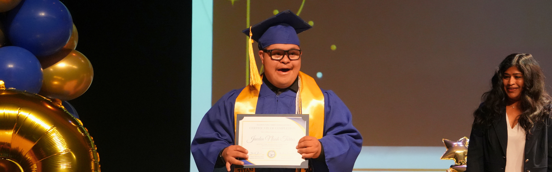 A student in a blue graduation gown holding a diploma, celebrating on stage.