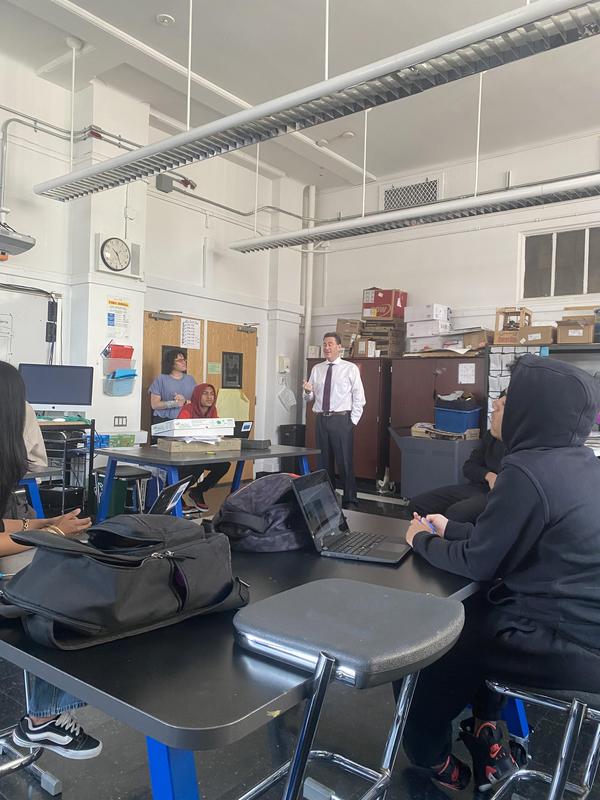 A teacher addresses a group of students in a classroom with laptops and various supplies.