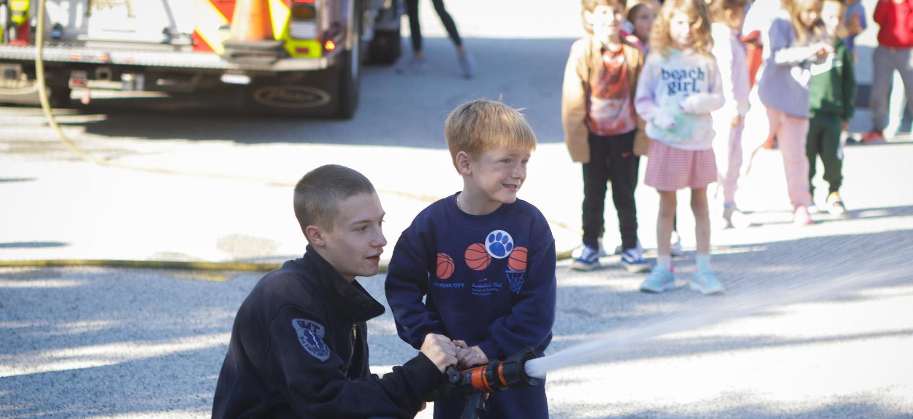 A child and a young adult spraying water from a fire hose at an outdoor event.