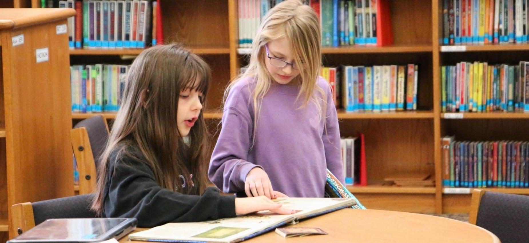 Two girls examine a book together at a library table.