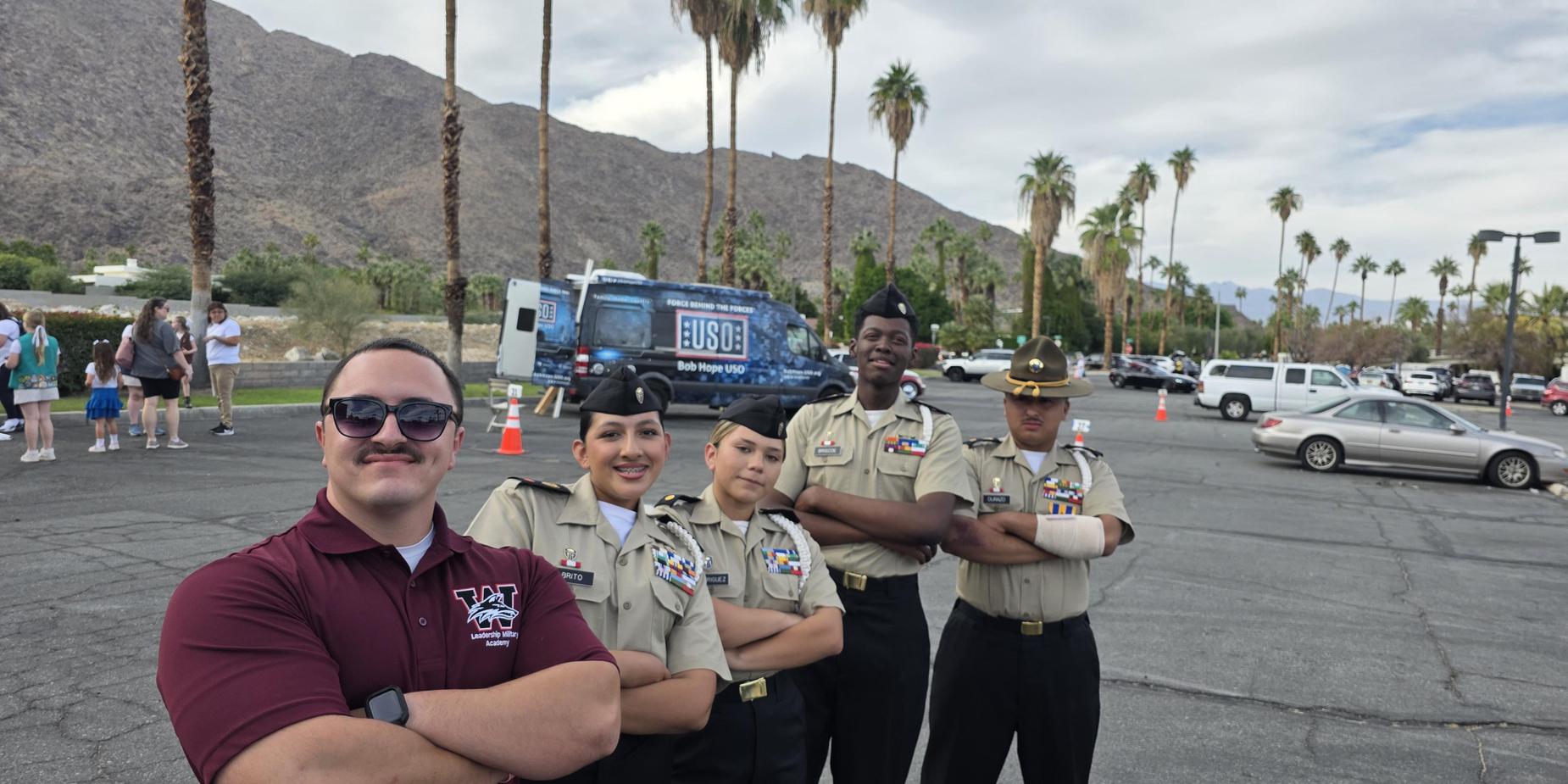 Five uniformed individuals posing together with palm trees and mountains in the background.