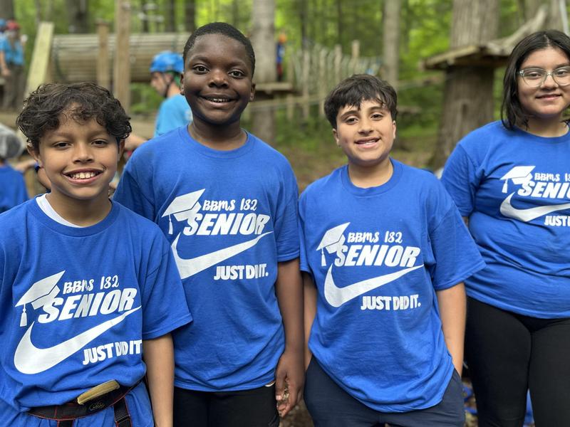 Four children in blue shirts with 'SENIOR' printed on them, smiling outdoors.