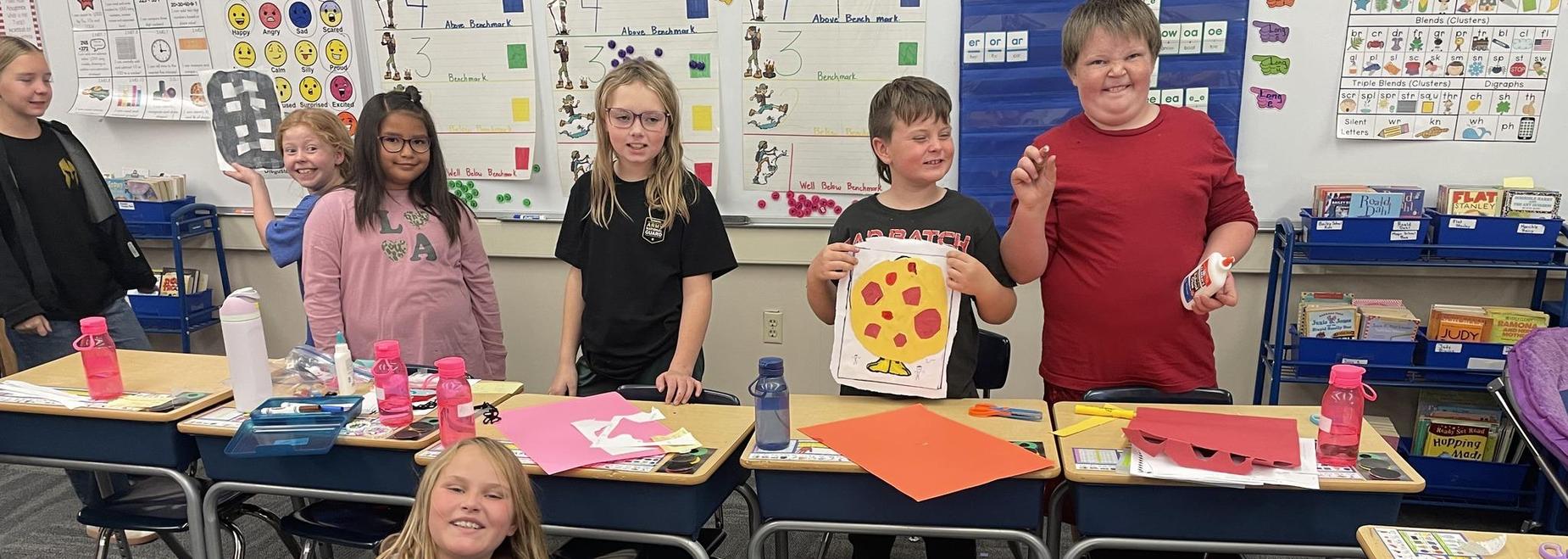 A group of students posing with their art projects at desks in a classroom.