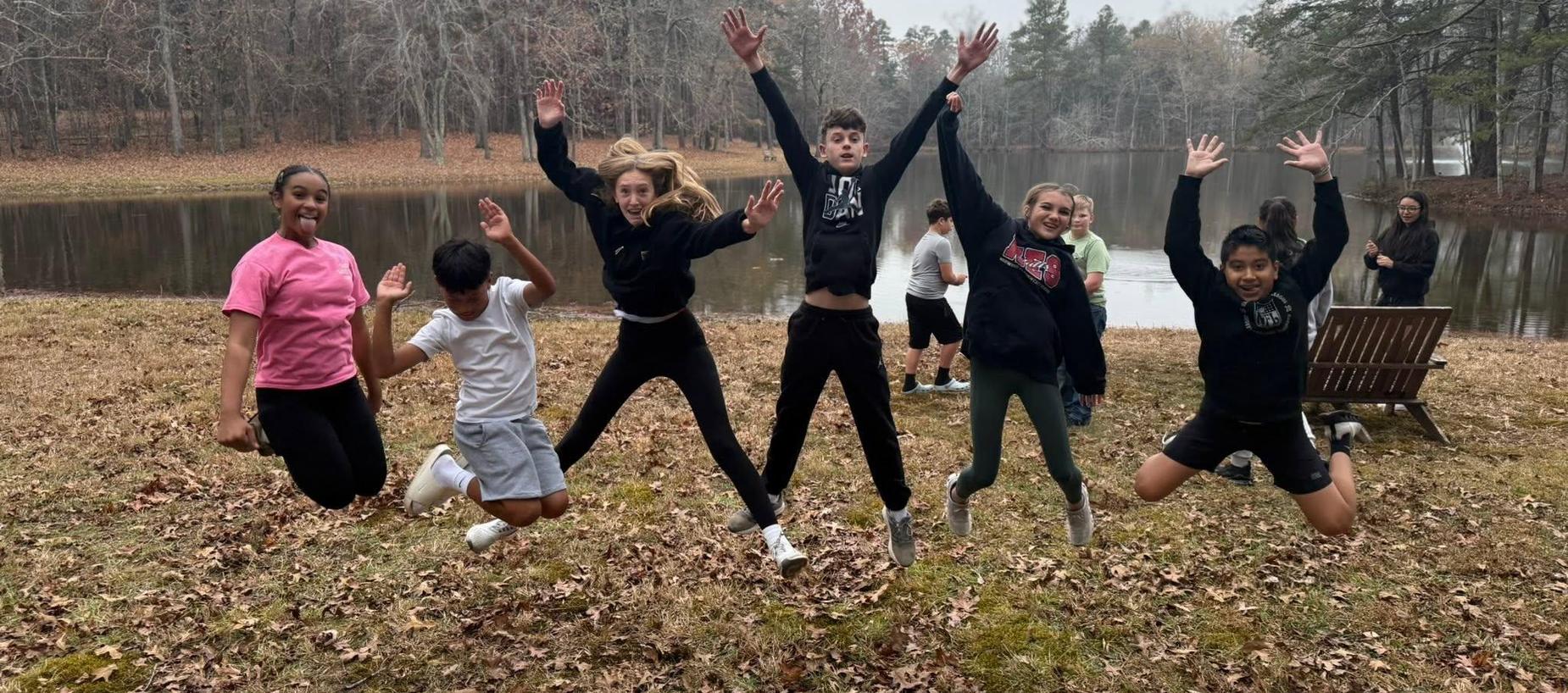 Group of six people jumping in the air by a lake in a wooded area.
