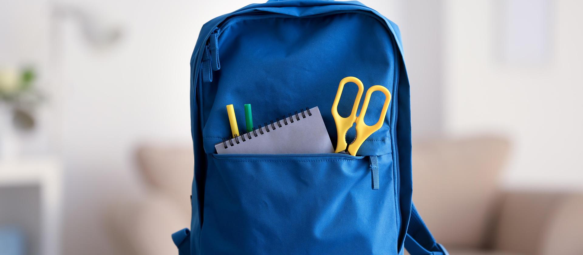 A blue backpack with scissors, a notepad, and colored pens in the front pocket.