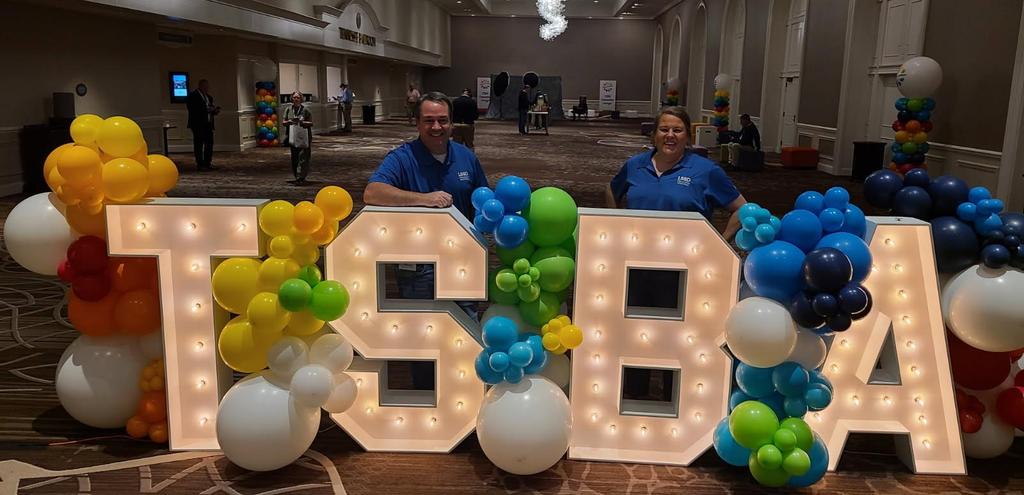 Lighted Large Letters spelling out TSBA with balloons around them and 2 Board Members standing behind