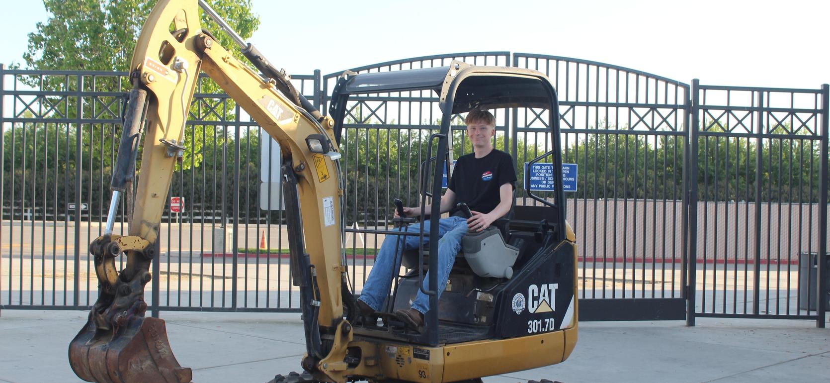 A person operating a yellow excavator outside a gated area.