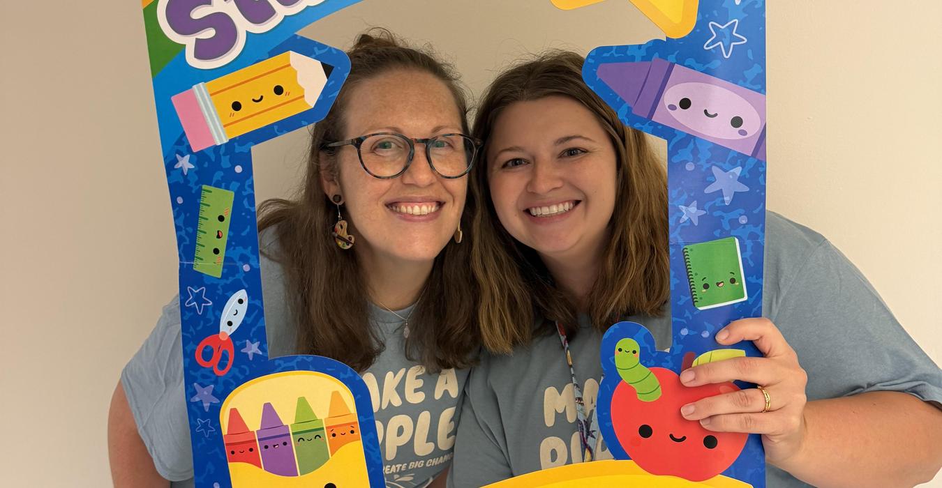Two smiling women holding a colorful frame decorated with school-themed graphics.