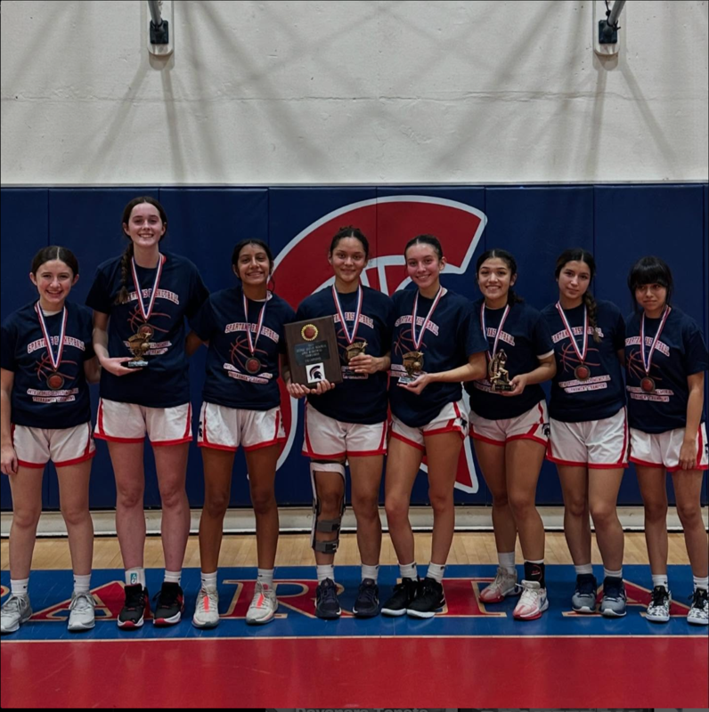 Eight girls’ basketball players in matching uniforms stand shoulder to shoulder on a gym court, smiling proudly with trophies and medals. One player holds a championship plaque, and several others hold individual awards. Behind them is a Spartan logo on the gym wall.