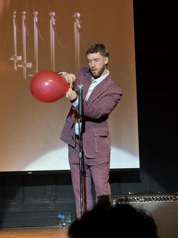 Performer in a suit inflating a red balloon on stage.