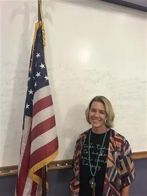 Woman smiling beside an American flag in front of a whiteboard.