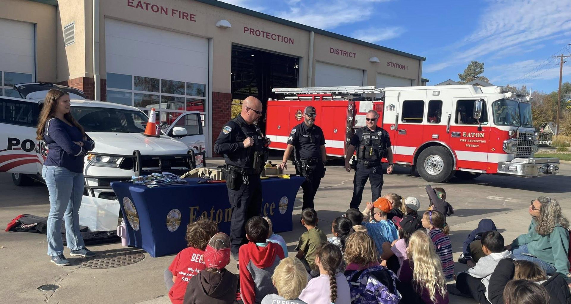 Children sitting on the ground watching police officers at an event outside a fire station.
