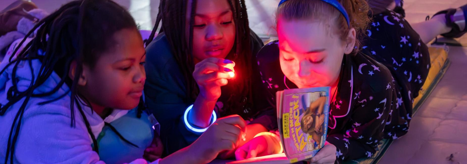 Three children reading a book together, illuminated by a soft light.