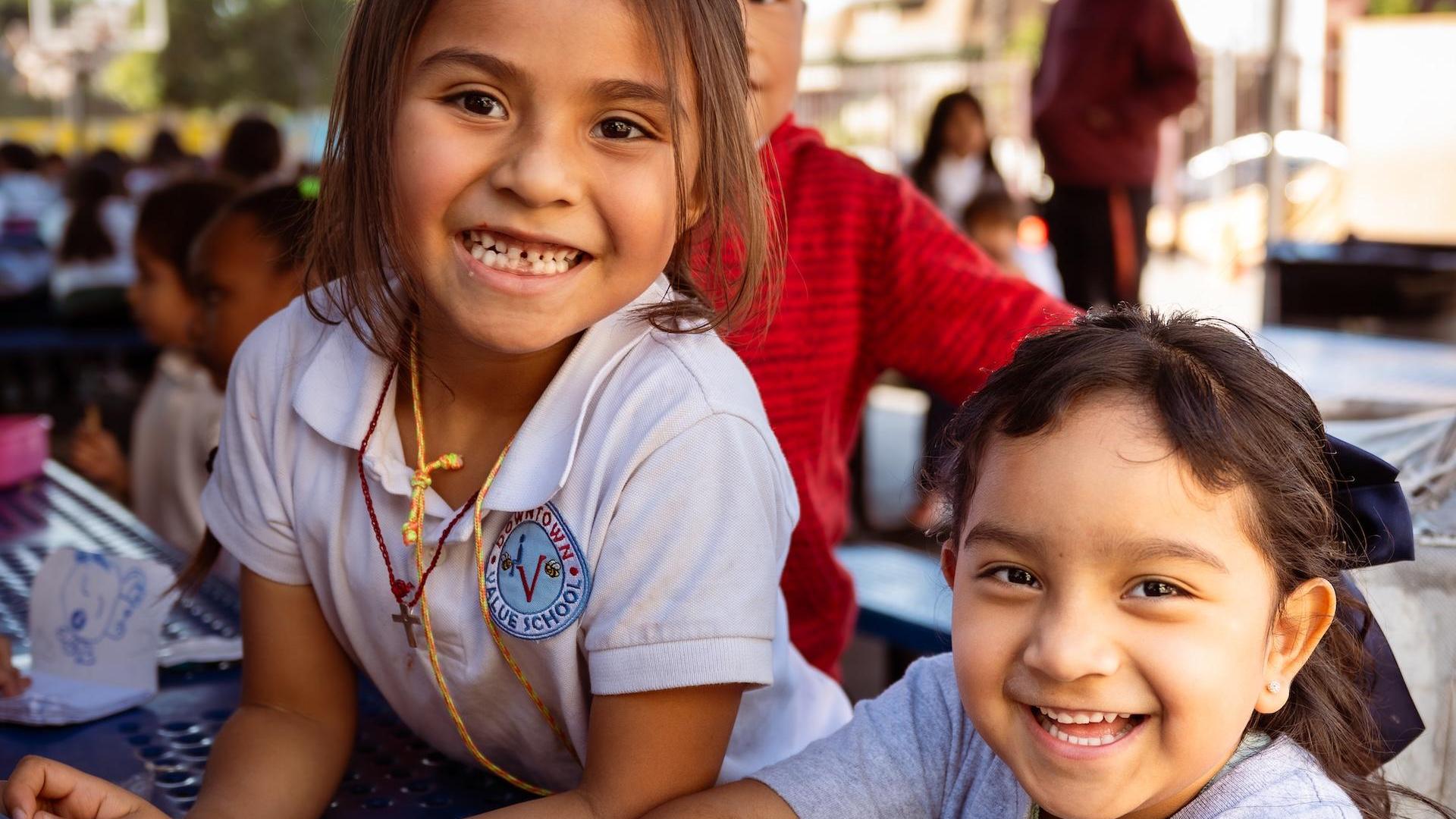 A smiling young girl sits at a table with classmates, enjoying their time together.