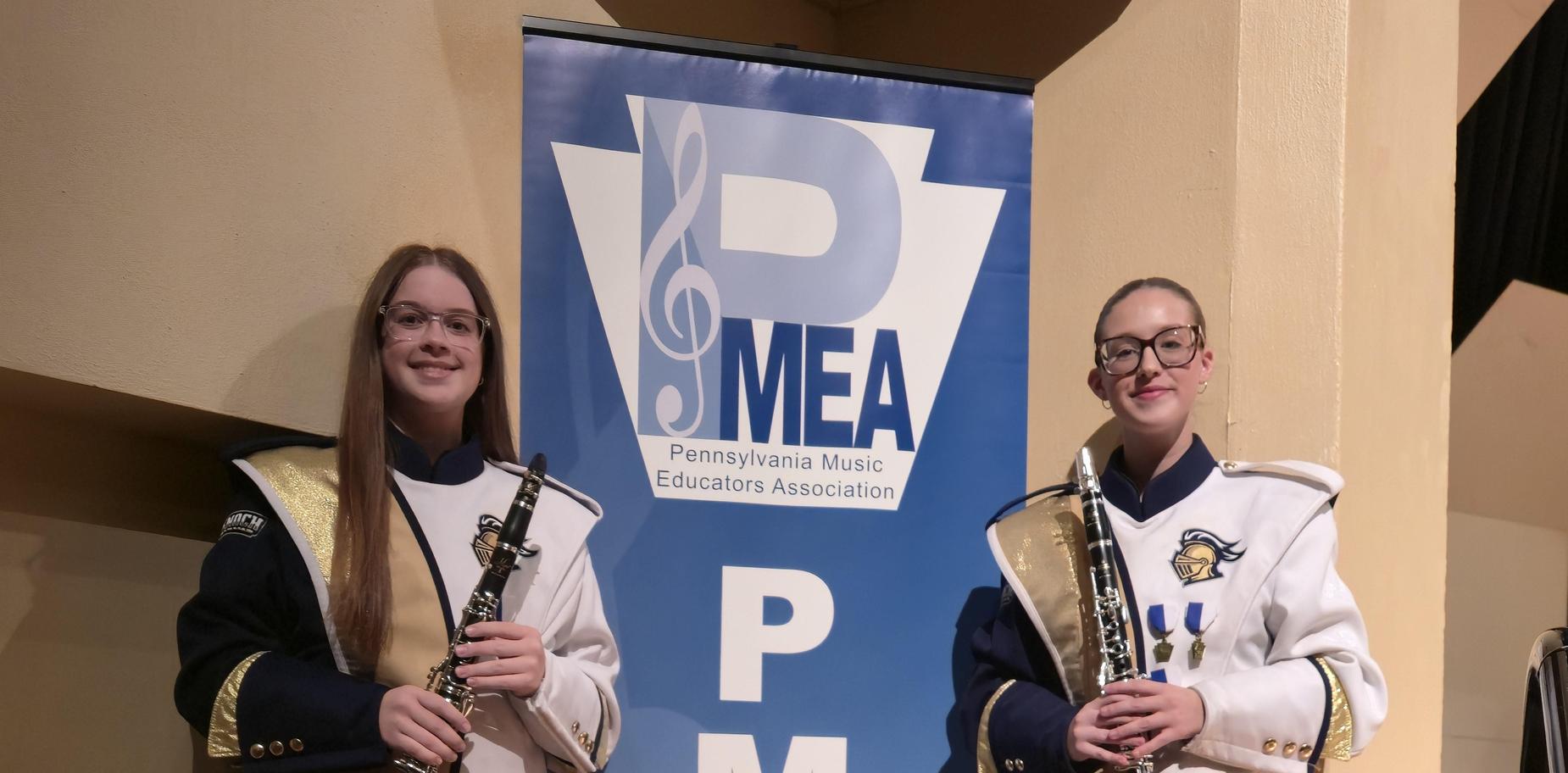 Two musicians in formal attire holding clarinets beside a PMEA banner.